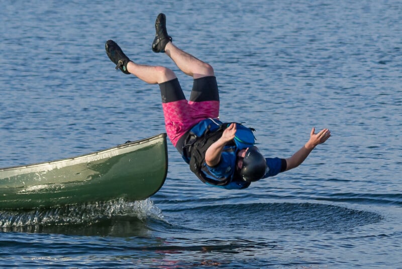 A person wearing a helmet and life vest falls backwards out of a green canoe into the water, with their legs in the air and arms outstretched. The canoe tilts from the motion, and water splashes around.