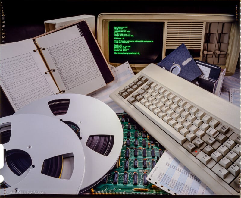 A vintage desktop computer with a green text screen, keyboard, floppy disks, open binder with data tables, punched cards, magnetic tape reels, and a circuit board, representing old data storage and computing technology.