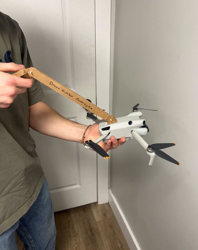 Un hombre sostiene un dron blanco mientras usa herramientas de cartón caseras etiquetadas "Regulador de cardán para drones" Ajusta el cardán del dron. El hombre está parado cerca de la puerta sobre paredes blancas y suelo de madera.