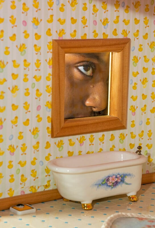 Primer plano de un rostro humano reflejado en un pequeño espejo encima de una mini bañera con decoración floral y papel pintado con una ilustración de un pato amarillo de fondo.