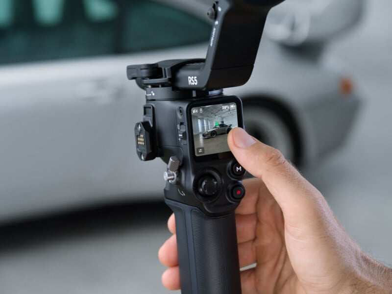 A hand holds a camera gimbal with a screen displaying a car being filmed. The blurred background shows part of a gray car. The gimbal is labeled "RS3.