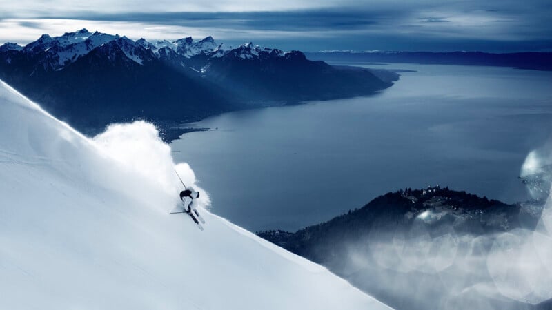 A skier carves down a snowy mountain slope, creating a spray of powder, with a large lake and snow-capped mountains visible in the distant background under a cloudy sky.