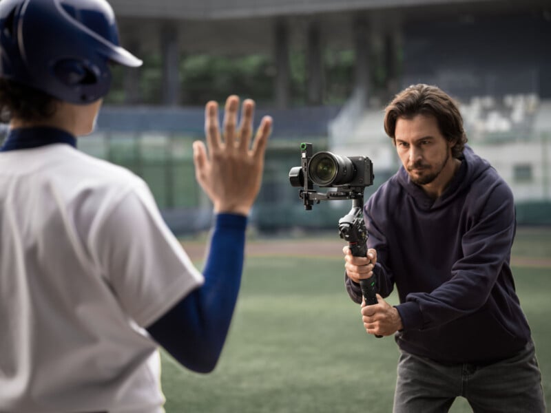 A man holding a camera on a stabilizer films a baseball player, who is raising one hand, on an outdoor field. The focus is on the cameraman as he captures the player’s actions up close.