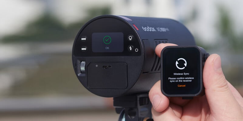 Una mano que sostiene un reloj inteligente que muestra un mensaje de sincronización inalámbrica cerca de la unidad de flash de una cámara profesional que muestra la pantalla de confirmación de sincronización. El fondo está borroso, lo que sugiere un escenario al aire libre.