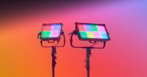 Two professional LED panel lights on stands emit multicolored light patterns against a vibrant gradient background of orange, pink, and purple hues.
