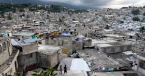 A densely packed hillside cityscape with concrete buildings and makeshift structures covered in tarps, showing signs of crowding and poverty under a cloudy sky, with mountains visible in the background.