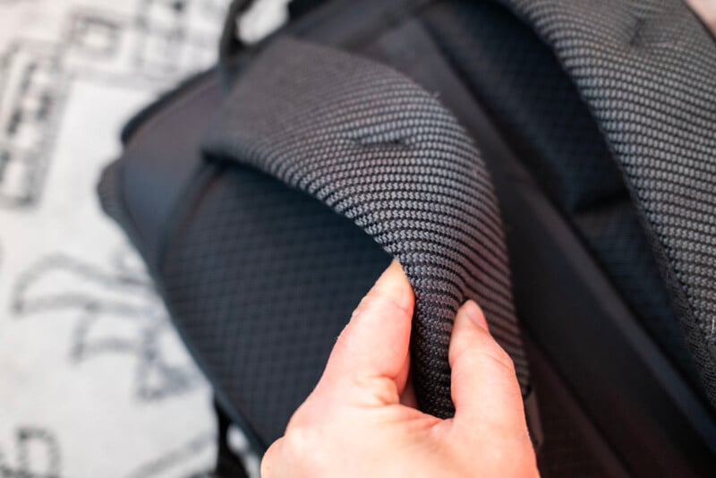 A close-up of a hand pinching the padded, textured strap of a dark-colored backpack, showing the thick, woven material and the padding inside. The background is blurred with faint patterns visible.