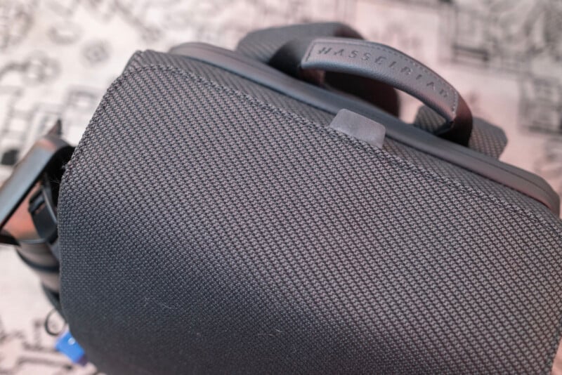 A close-up photo of a textured black bag with a handle, featuring the brand name "Masserfakar" embossed on a leather tag. The background is blurred and patterned in light colors.