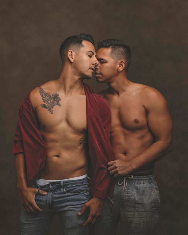 Two shirtless men stand close together, facing each other intimately. One wears an open red shirt and jeans, showing a large eagle tattoo on his chest. The other wears light pants. They appear to be about to kiss.