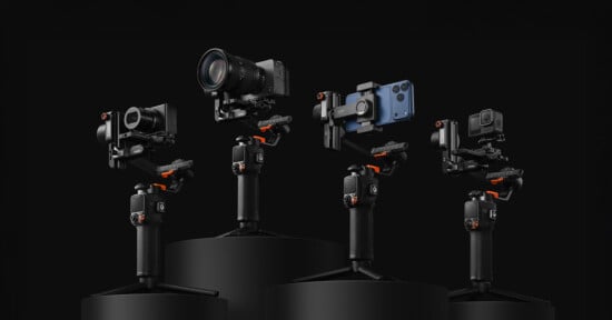 Four camera stabilizers, each holding a different device: a DSLR camera, a mirrorless camera, a smartphone, and an action camera, are displayed on black podiums against a dark background.