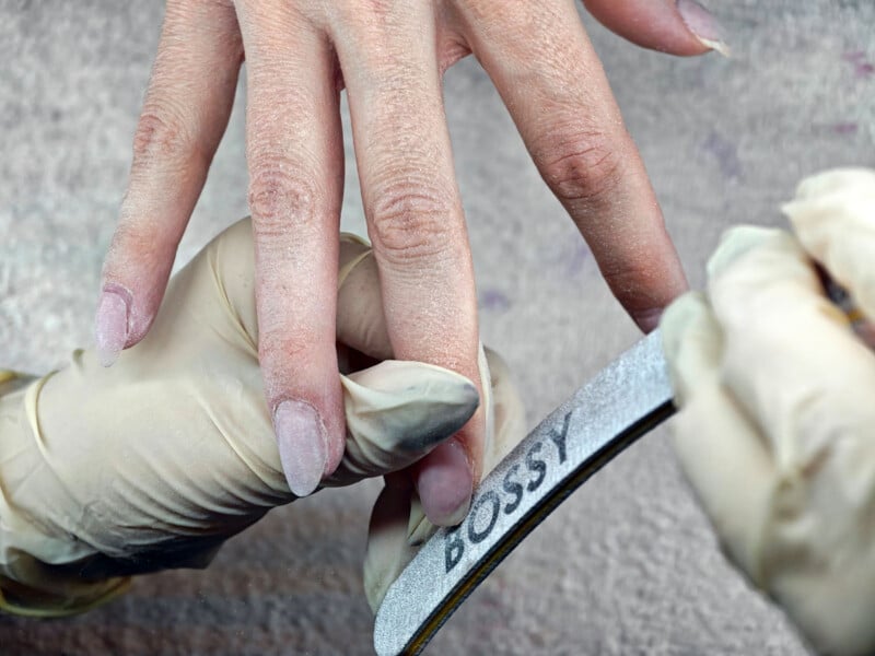 A gloved hand files a person's fingernails using a nail file labeled "BOSSY." Nail dust is visible on the fingers and gloves, indicating an ongoing manicure process.