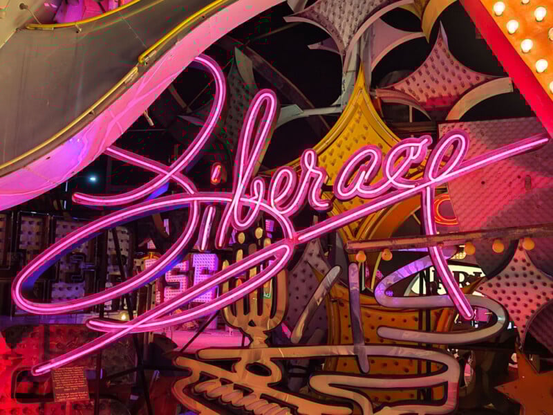 A bright pink neon sign spells "Liberace" in cursive letters, surrounded by colorful, retro-style lights and decorative shapes in a vintage, nighttime setting.