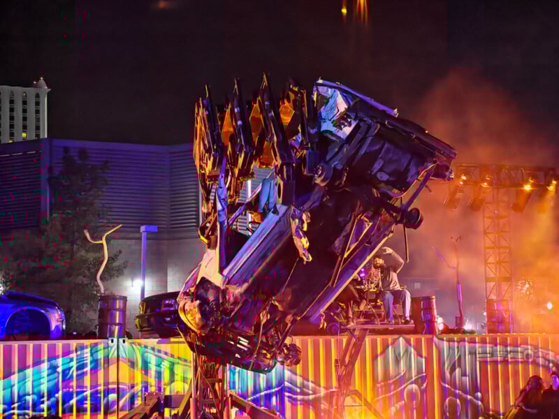 A car is lifted vertically by a giant mechanical claw at night, surrounded by colorful lights, smoke, and a vibrant, illuminated backdrop at an outdoor event or show.