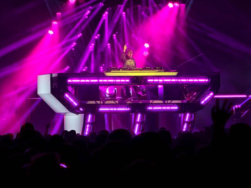 A DJ stands on a futuristic, illuminated stage with bright purple and pink lights, performing in front of a large, cheering crowd during a night concert.