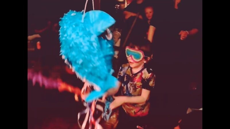 A child wearing a blindfold and colorful shirt swings a stick at a blue shark-shaped piñata while people watch in the background at a party.