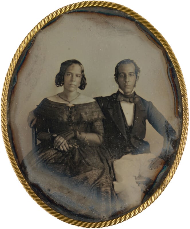 A vintage oval photograph in a gold frame shows a seated woman in a dark dress and gloves beside a man in a suit and bow tie, both looking at the camera with neutral expressions.