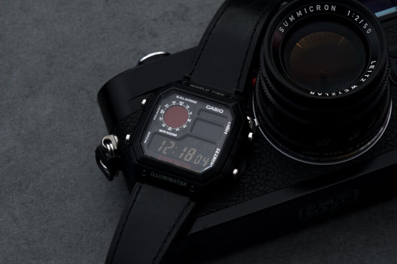 A black Casio digital wristwatch with a world time feature rests on top of a vintage black camera with a large lens, all set against a dark gray surface.