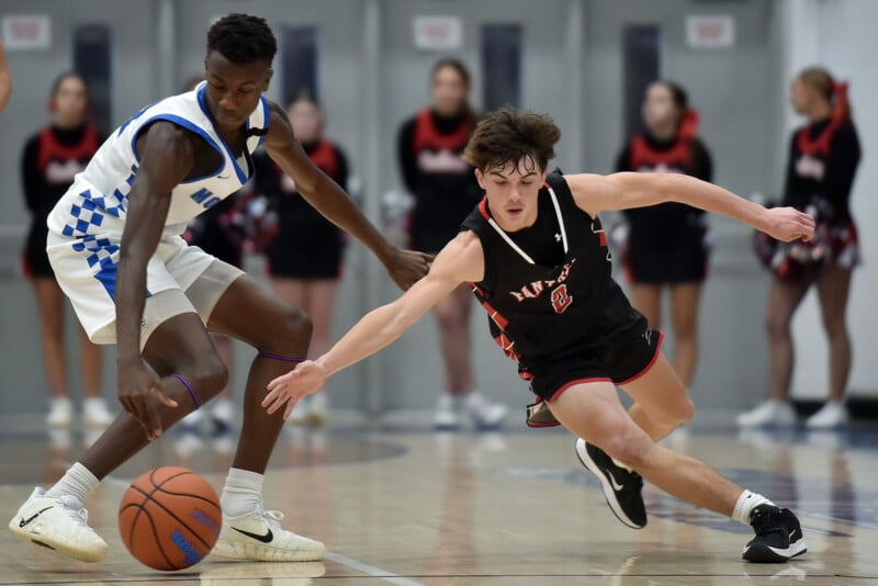 En la cancha de gimnasia, un jugador de baloncesto con uniforme blanco y azul dribla una pelota mientras otro jugador con uniforme negro y rojo lucha por alcanzar la pelota, con porristas borrosas en el fondo.