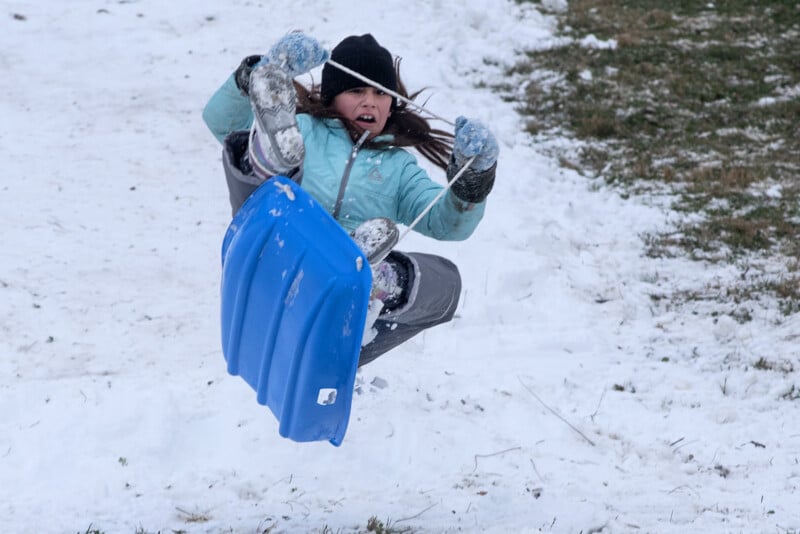 Una joven sentada en un trineo azul, vestida con una chaqueta verde azulado, sombrero negro y guantes, parece sorprendida mientras rebota por una montaña nevada en el aire, con parches de hierba visibles en el fondo.