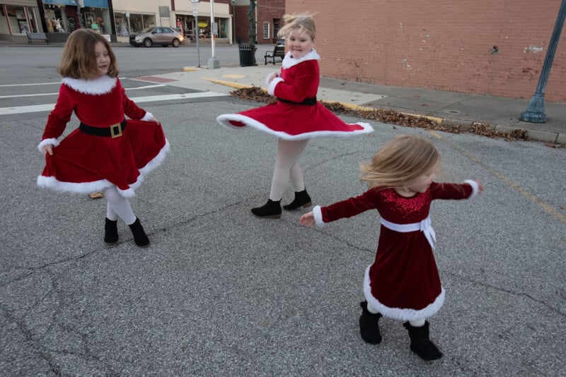 Tres niñas con vestidos rojos de fiesta daban vueltas y jugaban en un estacionamiento vacío. Las chicas llevaban medias blancas y botas negras, y dos de los vestidos tenían ribetes de piel blanca. Al fondo se ven claramente edificios y tiendas.