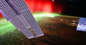 View from space showing the edge of a solar panel and part of the International Space Station with a vivid red and green aurora over the Earth’s surface, and city lights visible below.