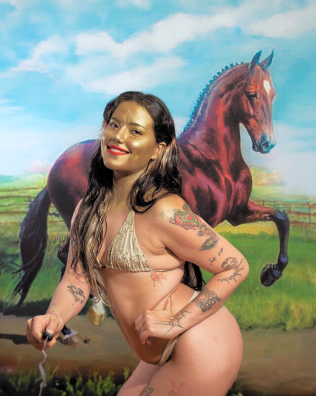 Un hombre sonriente con el pelo largo, pintura facial rubia y varios tatuajes en bikini se encuentra frente a una pintura de un caballo marrón galopando en un prado bajo un cielo azul y nubes blancas.