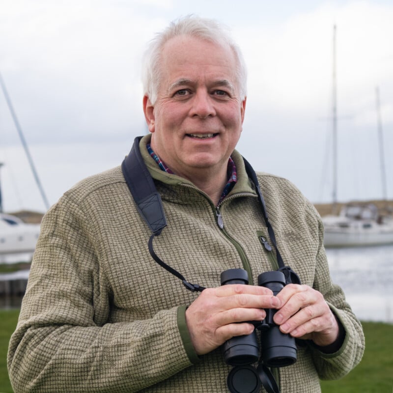 An older man with gray hair smiles outdoors, holding binoculars. He wears a green zip-up sweater, standing near water with sailboats and a cloudy sky in the background.