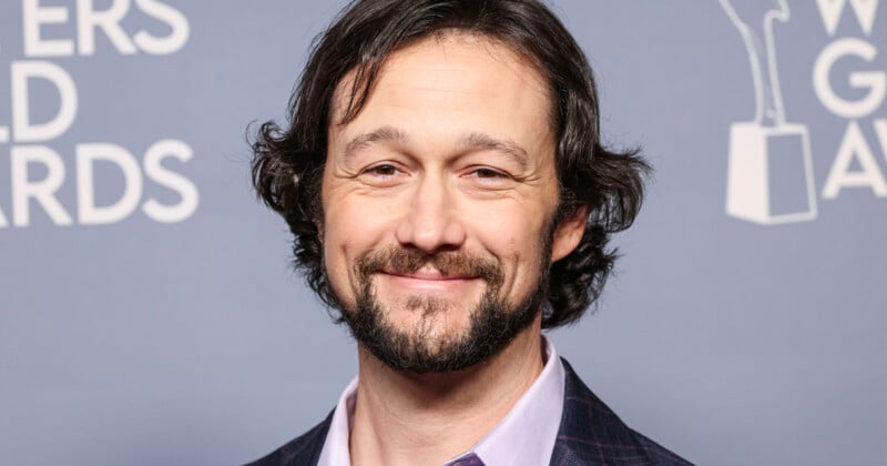 Un hombre con cabello negro ondulado y barba sonríe frente a un fondo gris con texto blanco y un patrón de estatuilla de premio. Llevaba una camisa con cuello rosa claro y una chaqueta a cuadros.