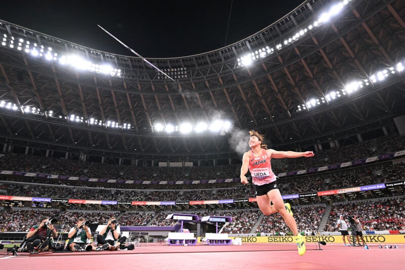 A female javelin thrower in mid-action on a stadium track, with the javelin airborne above her. Bright stadium lights, a large audience, and photographers capturing the event are visible in the background.