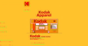 Illustration of a Kodak storefront labeled "Kodak Apparel" with shirts and film in the window, set on a yellow background. The Kodak logo appears at the top left and above the store. Text reads: "You Press the Button, We do the Rest.