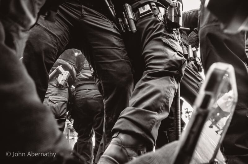 A black-and-white photo showing a close-up of law enforcement officers standing together, with visible holstered weapons and tactical gear. One person appears to be holding up a phone in the bottom right corner.