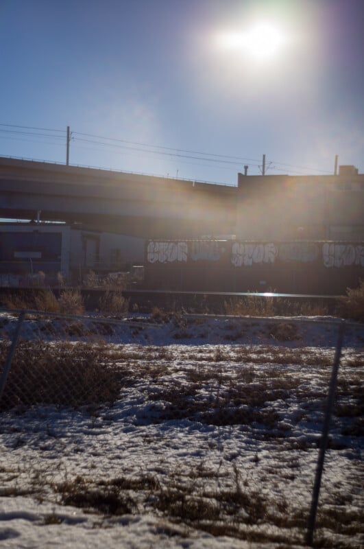 A sunlit urban scene with a chain-link fence, snow-covered ground, dried grass, and graffiti-covered buildings and train cars beneath an overpass, with the sun shining brightly in the blue sky.