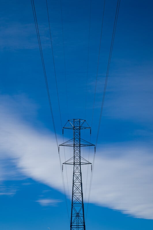 A tall electrical transmission tower stands against a bright blue sky with wispy white clouds, with multiple power lines extending outward from the top.