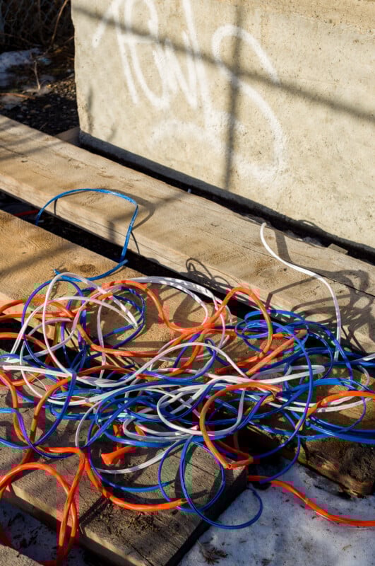 A tangle of red, blue, and white cables lies on wooden boards outdoors, next to a concrete wall with faded graffiti under sunlight.