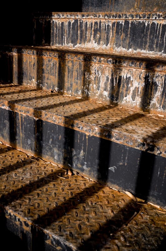 Close-up of rusty, metal stairs with textured surfaces and signs of corrosion. Light casts strong shadows from a railing across the steps, highlighting the rough and weathered condition.
