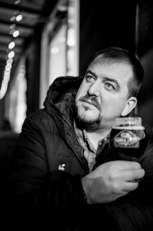 A man with a beard and mustache, wearing a jacket, sits indoors holding a glass of dark beer. He gazes thoughtfully upward. The image is in black and white, with soft, blurred lights in the background.