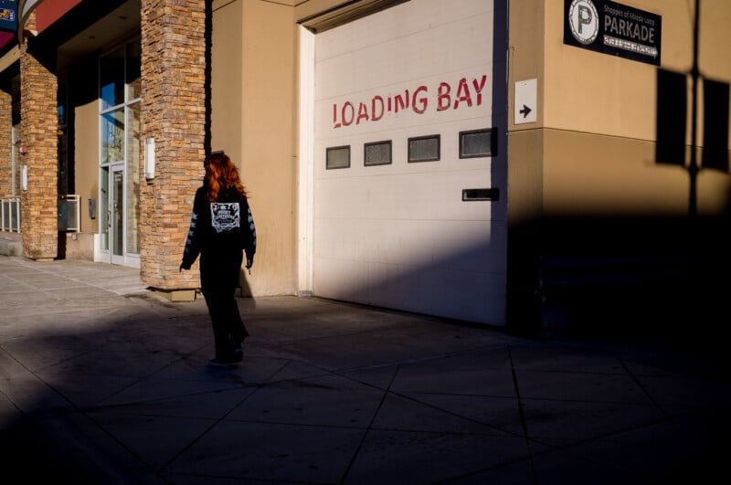 A person with long red hair walks past a white garage door labeled "LOADING BAY" on a city sidewalk, with sunlight casting sharp shadows on the building.