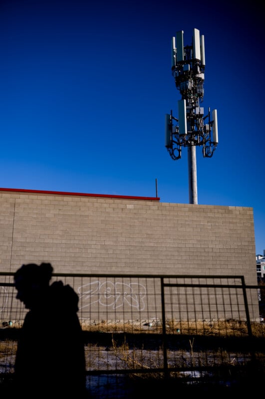 A person’s shadow is cast on a fence in front of a beige brick building with graffiti, with a tall cell tower extending above the building against a clear blue sky.