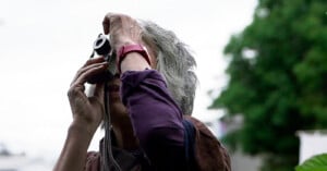 A person with gray hair, wearing a red wristwatch, holds a camera up to their face and looks through the viewfinder outdoors, with blurred greenery in the background.