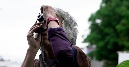 A person with gray hair, wearing a red wristwatch, holds a camera up to their face and looks through the viewfinder outdoors, with blurred greenery in the background.