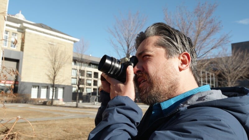 A man with gray hair and a beard wears a blue jacket and holds a camera up to his eye, photographing outdoors on a sunny day with trees and buildings in the background.