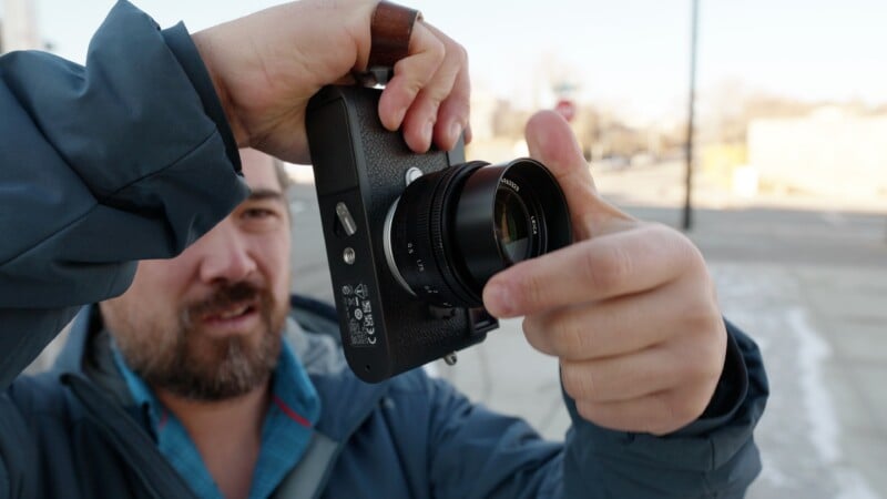 A person holding a camera close to their face, adjusting the lens and preparing to take a photo outdoors on a bright day. The background is slightly blurred, showing a sidewalk and some buildings.