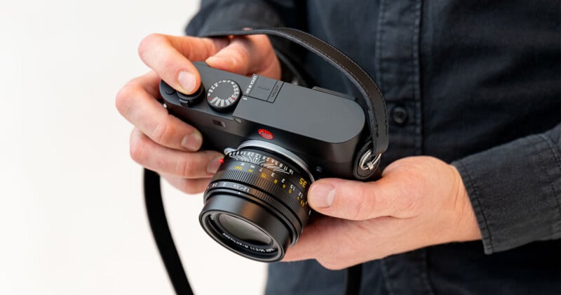 A person in a dark shirt holds a black Leica camera with both hands, showing the camera’s lens, dials, and leather strap against a light background.