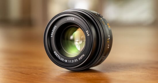 A Leica camera lens, specifically a Noctilux-M 50mm f/1.2 ASPH, rests on a wooden surface with soft, blurred background lighting.