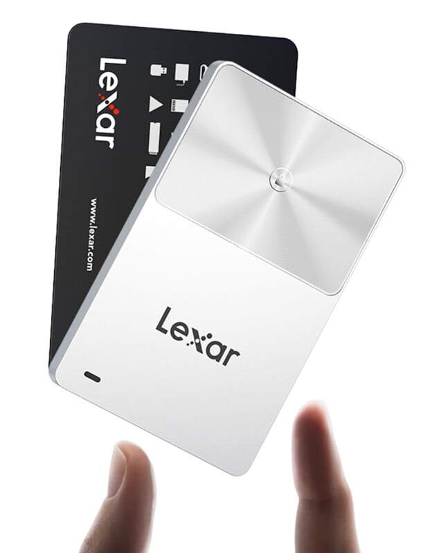 A hand reaches for a compact Lexar portable solid state drive, which is shown in front of a black card with the Lexar logo and website. The SSD is silver with a modern, sleek design.