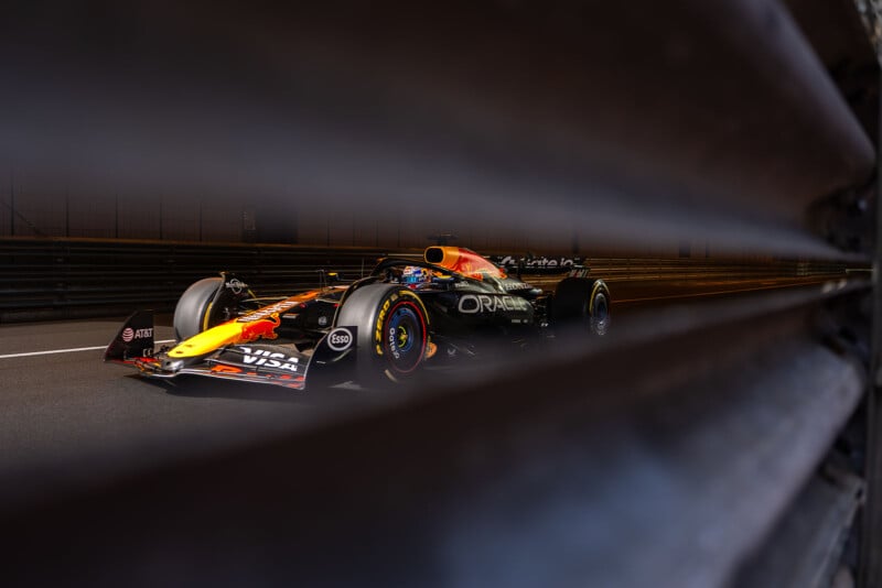 A Red Bull Formula 1 car is seen racing on a track, photographed through the slats of a metal barrier, creating a dramatic, partially obstructed view.