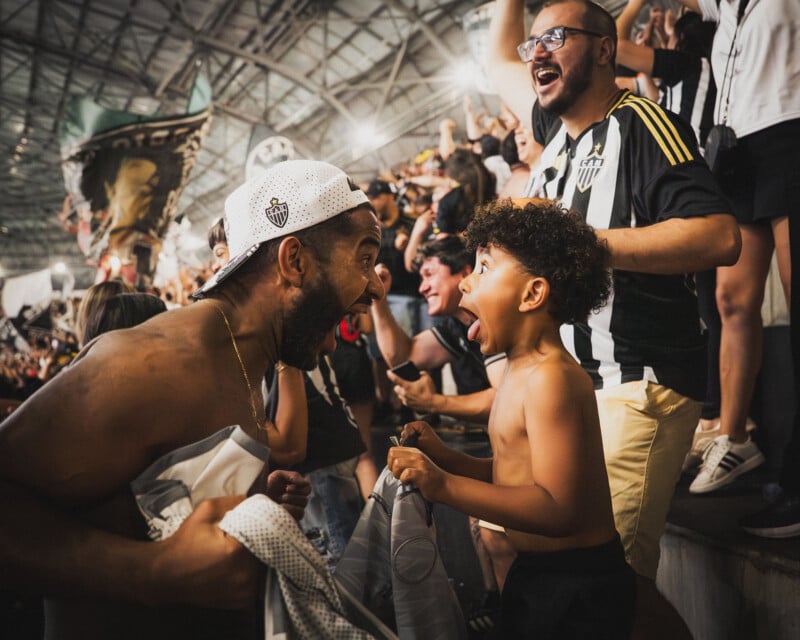 A shirtless man and a young boy excitedly yell at each other in celebration at a crowded stadium, surrounded by cheering soccer fans wearing black and white jerseys.