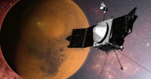 A spacecraft with solar panels orbits close to the reddish surface of Mars, set against a backdrop of space filled with stars and galaxies.