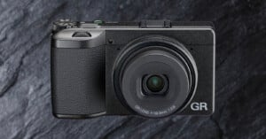 A compact black digital camera with a textured grip and a prominent lens, labeled "GR," is shown against a dark, stone-textured background.