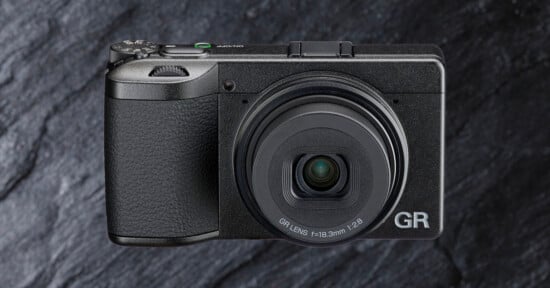A compact black digital camera with a textured grip and a prominent lens, labeled "GR," is shown against a dark, stone-textured background.